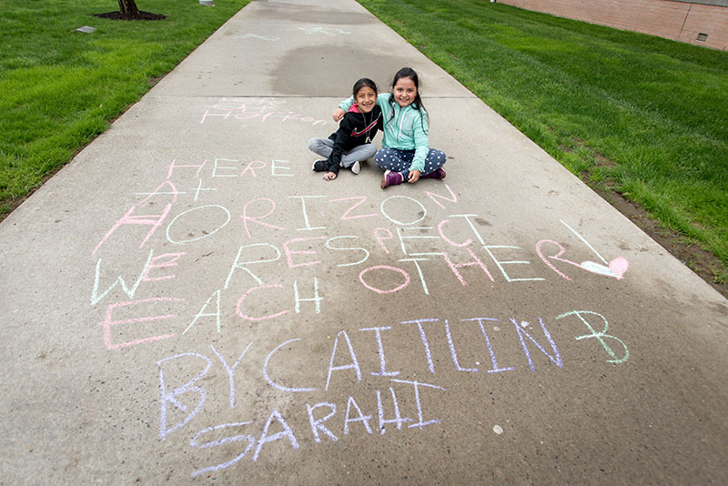 Two young girls sitting on sidewalk with 'Here at Horizons We Respect Each Other' written in chalk